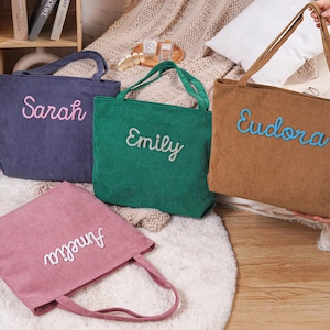 May include: Four corduroy tote bags in shades of blue, green, brown, and pink. Each bag features a name embroidered in a contrasting color: Sarah, Emily, Eudora, and Amelia. The bags have matching handles.