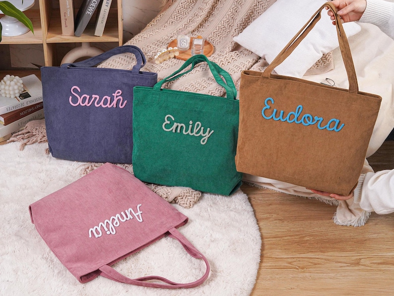 May include: Four corduroy tote bags in shades of blue, green, brown, and pink. Each bag features a name embroidered in a contrasting color: Sarah, Emily, Eudora, and Amelia. The bags have matching handles.