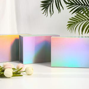May include: Three rectangular gift boxes with iridescent, color-shifting surfaces. The boxes are arranged in a row, with a floral arrangement in front. The boxes reflect a range of colors, including pink, yellow, and blue.