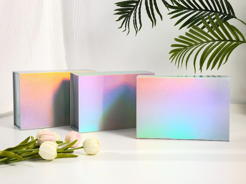 K&ouml;nnte beinhalten: Drei rechteckige Geschenkboxen mit schillernden, farbver&auml;ndernden Oberfl&auml;chen. Die Boxen sind in einer Reihe angeordnet, mit ein paar Kunstblumen und Gr&uuml;nzeug davor. Die Boxen reflektieren eine Reihe von Farben, darunter Rosa, Gelb und Blau.