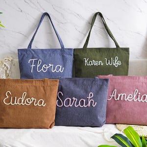 May include: Five personalized tote bags in various colors: denim blue, olive green, brown, dark blue, and dusty rose. Each bag features a name embroidered in a cursive font. The bags are displayed on a white surface.