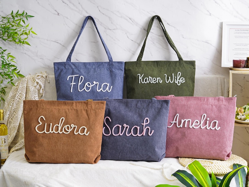 May include: Five personalized tote bags in various colors: denim blue, olive green, brown, dark blue, and dusty rose. Each bag features a name embroidered in a cursive font. The bags are displayed on a white surface.