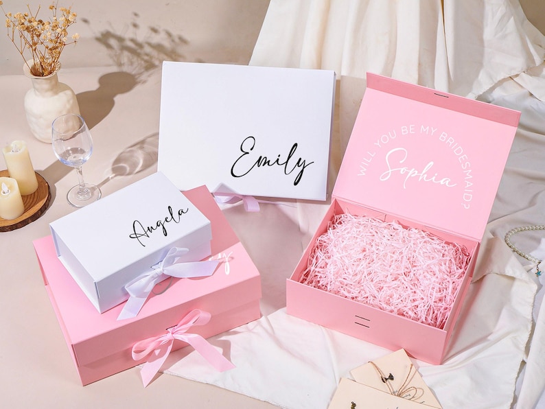Pu&ograve; includere: Scatole regalo bianche e rosa, con nomi come "Emily" e "Angela" in corsivo. Una scatola &egrave; aperta, riempita di carta rosa, e recita "Will you be my Bridesmaid?" con il nome "Sophia".