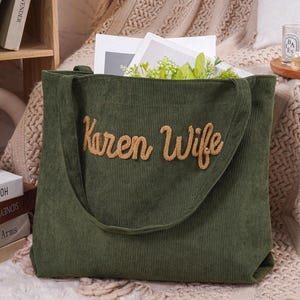 May include: A green corduroy tote bag with the words "Karen Wife" embroidered in a light brown, cursive font. The bag has a long shoulder strap and is filled with white paper and greenery.