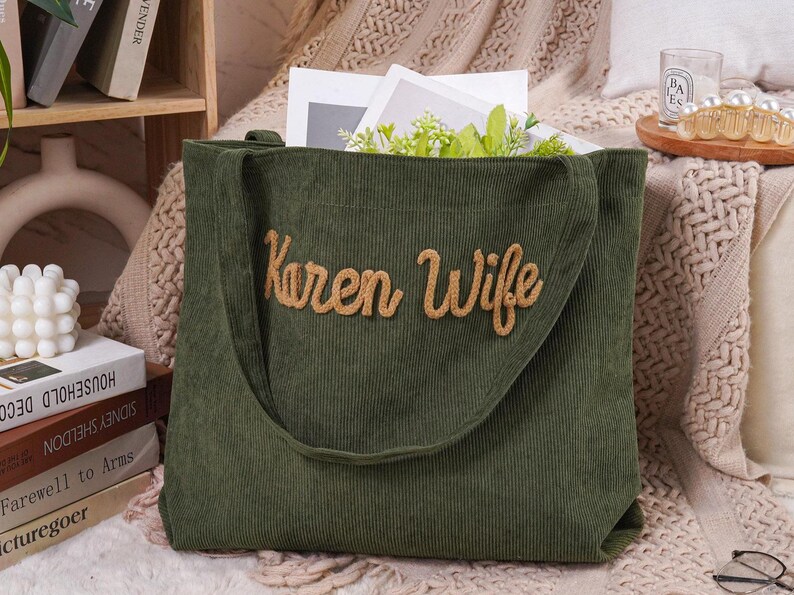 May include: A green corduroy tote bag with the words "Karen Wife" embroidered in a light brown, cursive font. The bag has a long shoulder strap and is filled with white paper and greenery.