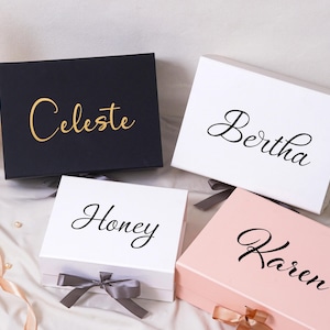 Pu&ograve; includere: Quattro scatole regalo rettangolari con coperchi incernierati e chiusure a nastro. Una scatola nera presenta il testo dorato Celeste, due scatole bianche presentano il testo nero Bertha e Honey, una scatola rosa chiaro presenta il testo nero Karen.