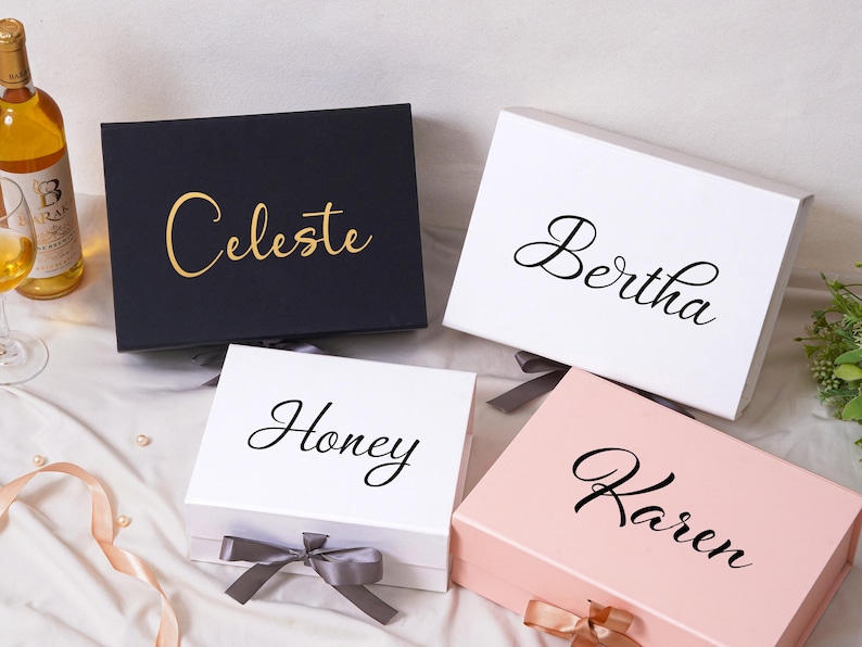 Pu&ograve; includere: Quattro scatole regalo rettangolari con coperchi incernierati e chiusure a nastro. Una scatola nera presenta il testo dorato Celeste, due scatole bianche presentano il testo nero Bertha e Honey, una scatola rosa chiaro presenta il testo nero Karen.