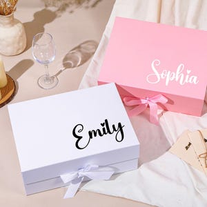 Pu&ograve; includere: Due scatole regalo, una bianca e una rosa, con nomi in corsivo. La scatola bianca ha "Emily" in corsivo nero e la scatola rosa ha "Sophia" in corsivo bianco. Entrambe le scatole hanno una chiusura a nastro. Un bicchiere da vino, candele e un vaso sono anche nell'immagine.