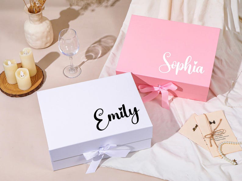 Pu&ograve; includere: Due scatole regalo, una bianca e una rosa, con nomi in corsivo. La scatola bianca ha "Emily" in corsivo nero e la scatola rosa ha "Sophia" in corsivo bianco. Entrambe le scatole hanno una chiusura a nastro. Un bicchiere da vino, candele e un vaso sono anche nell'immagine.
