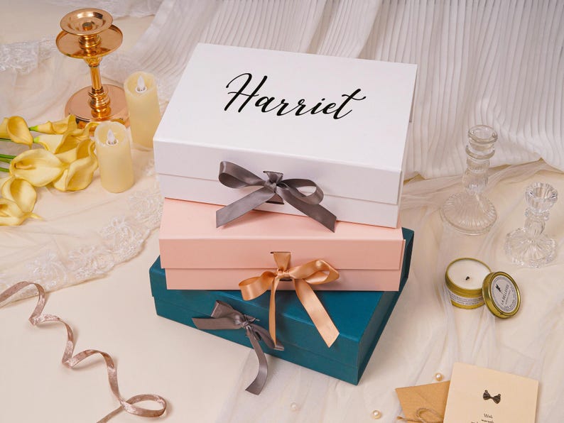 Pu&ograve; includere: Una pila di tre scatole regalo rettangolari nei colori bianco, rosa chiaro e verde acqua scuro. La scatola bianca superiore presenta il nome Harriet in corsivo nero. Ogni scatola &egrave; chiusa da un fiocco di raso rispettivamente grigio, oro e grigio.