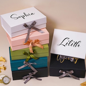 Pu&ograve; includere: Una pila di cinque scatole regalo rettangolari nei colori bianco, rosa chiaro, verde oliva, verde acqua e nero, ciascuna dotata di chiusura a nastro. Una scatola regalo nera separata con nastro &egrave; posizionata a destra. Le scatole bianche sono personalizzate con i nomi Sophia e Lilith in corsivo nero.