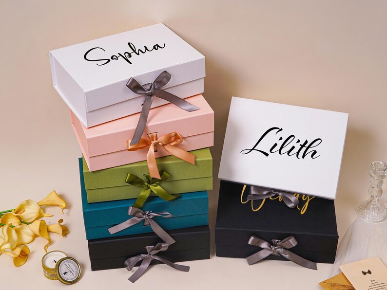 Pu&ograve; includere: Una pila di cinque scatole regalo rettangolari nei colori bianco, rosa chiaro, verde oliva, verde acqua e nero, ciascuna dotata di chiusura a nastro. Una scatola regalo nera separata con nastro &egrave; posizionata a destra. Le scatole bianche sono personalizzate con i nomi Sophia e Lilith in corsivo nero.
