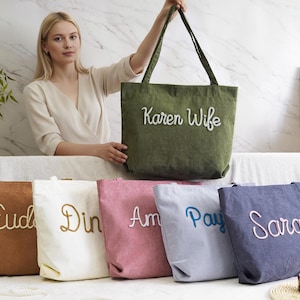 May include: A collection of canvas tote bags in various colors, each embroidered with a name or title. The bags are in shades of brown, white, pink, blue, and navy, with the text in complementary colors. One green bag reads "Karen Wife".