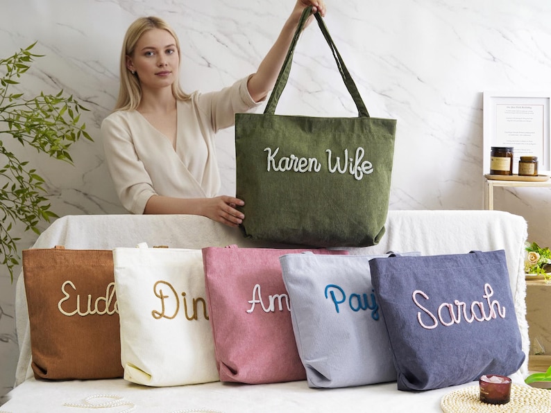 May include: A collection of canvas tote bags in various colors, each embroidered with a name or title. The bags are in shades of brown, white, pink, blue, and navy, with the text in complementary colors. One green bag reads "Karen Wife".