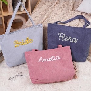 May include: Three corduroy tote bags in gray, navy blue, and pink. The gray bag has "Bride" in yellow script, the navy bag has "Flora" in white script, and the pink bag has "Amelia" in white script. The bags have handles.