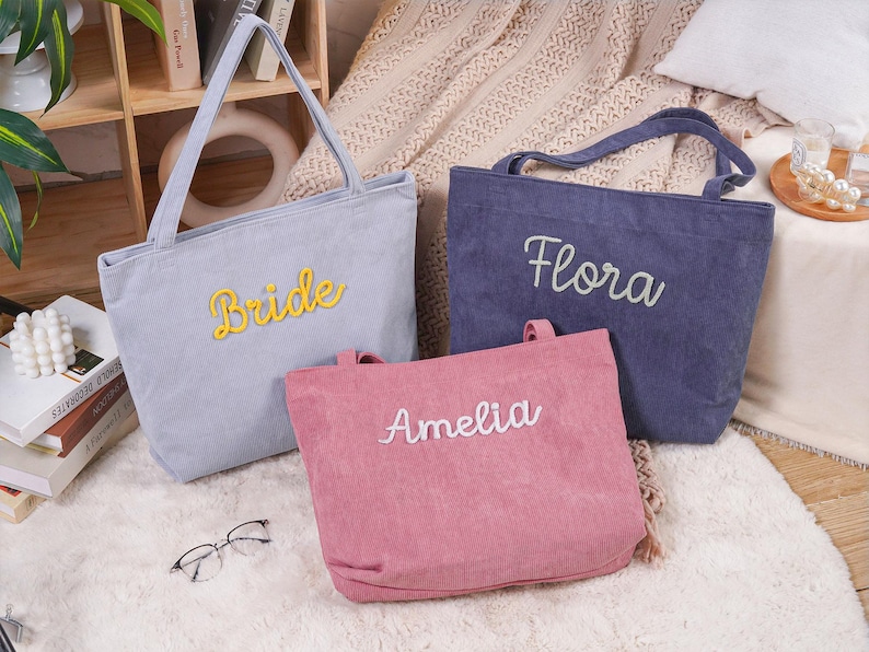 May include: Three corduroy tote bags in gray, navy blue, and pink. The gray bag has "Bride" in yellow script, the navy bag has "Flora" in white script, and the pink bag has "Amelia" in white script. The bags have handles.