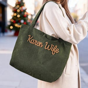 May include: Olive green corduroy tote bag with the words "Karen Wife" embroidered in a light brown script. The bag has a rectangular shape and a shoulder strap. The background is blurred, suggesting an outdoor setting.