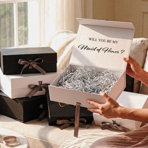Pu&ograve; includere: Una collezione di scatole regalo rettangolari bianche e nere, ciascuna con un nastro annodato. Una scatola bianca aperta contiene carta argentata sminuzzata e il testo "WILL YOU BE MY Maid of Honor ?" stampato all'interno del coperchio.