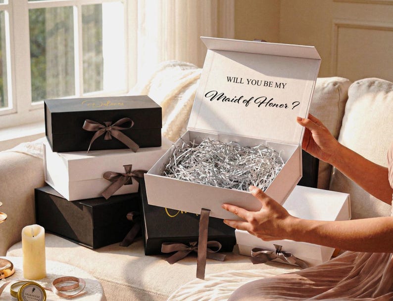 Pu&ograve; includere: Una collezione di scatole regalo rettangolari bianche e nere, ciascuna con un nastro annodato. Una scatola bianca aperta contiene carta argentata sminuzzata e il testo "WILL YOU BE MY Maid of Honor ?" stampato all'interno del coperchio.