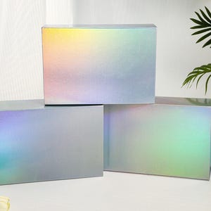 K&ouml;nnte beinhalten: Drei rechteckige Geschenkboxen mit holografischer, irisierender Oberfl&auml;che. Die Boxen sind gestapelt, wobei die obere Box einen Farbverlauf von Gelb, Rosa und Blau aufweist. Die anderen Boxen haben einen &auml;hnlichen Farbwechseleffekt mit gr&uuml;nen und blauen Farbt&ouml;nen.