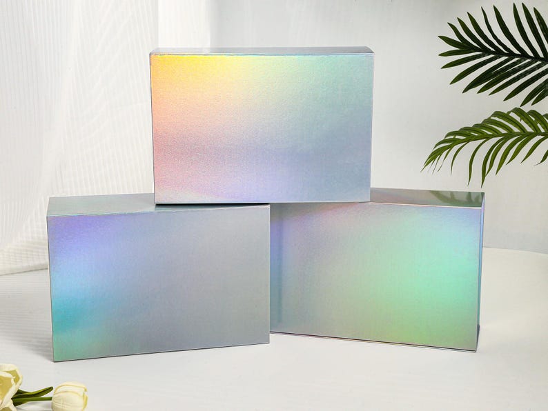 K&ouml;nnte beinhalten: Drei rechteckige Geschenkboxen mit holografischer, irisierender Oberfl&auml;che. Die Boxen sind gestapelt, wobei die obere Box einen Farbverlauf von Gelb, Rosa und Blau aufweist. Die anderen Boxen haben einen &auml;hnlichen Farbwechseleffekt mit gr&uuml;nen und blauen Farbt&ouml;nen.