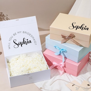 Pu&ograve; includere: Scatola regalo bianca con la scritta "Will you be my bridesmaid?" e il nome "Sophia". Scatole impilate in colori pastello: beige, azzurro e rosa. Ogni scatola ha un nastro. Le scatole sono piene di carta.