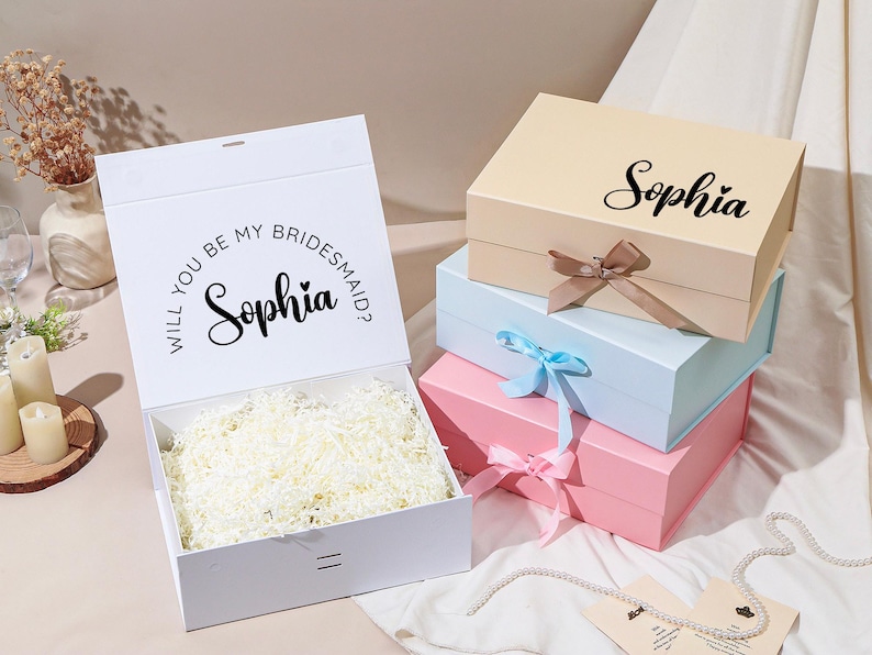 Pu&ograve; includere: Scatola regalo bianca con la scritta "Will you be my bridesmaid?" e il nome "Sophia". Scatole impilate in colori pastello: beige, azzurro e rosa. Ogni scatola ha un nastro. Le scatole sono piene di carta.