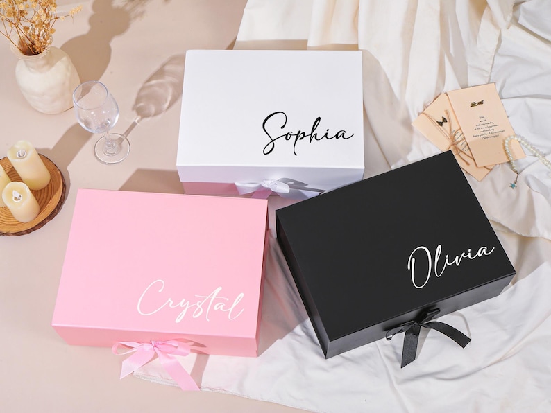 Pu&ograve; includere: Tre scatole regalo bianche, rosa e nere, ciascuna con un nome in elegante scrittura: Sophia, Crystal e Olivia. Le scatole hanno chiusure a nastro abbinate. Sono visibili anche un calice, candele e un piccolo biglietto.