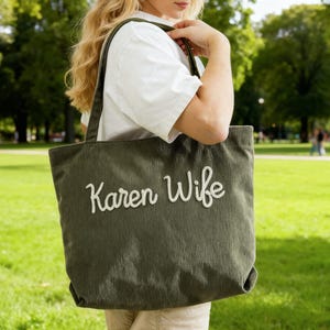May include: Olive green corduroy tote bag with the embroidered text "Karen Wife" in white. The bag has a rectangular shape and a wide shoulder strap. The background is a green park.