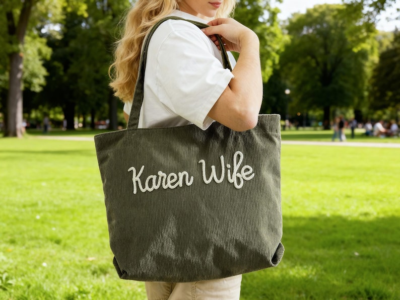 May include: Olive green corduroy tote bag with the embroidered text "Karen Wife" in white. The bag has a rectangular shape and a wide shoulder strap. The background is a green park.