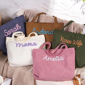 May include: A collection of corduroy tote bags in various colors, including blue, brown, white, green, and pink. Each bag features a different name embroidered in a contrasting color: Sarah, Eudora, Mama, Amelia, and Karen Wife.