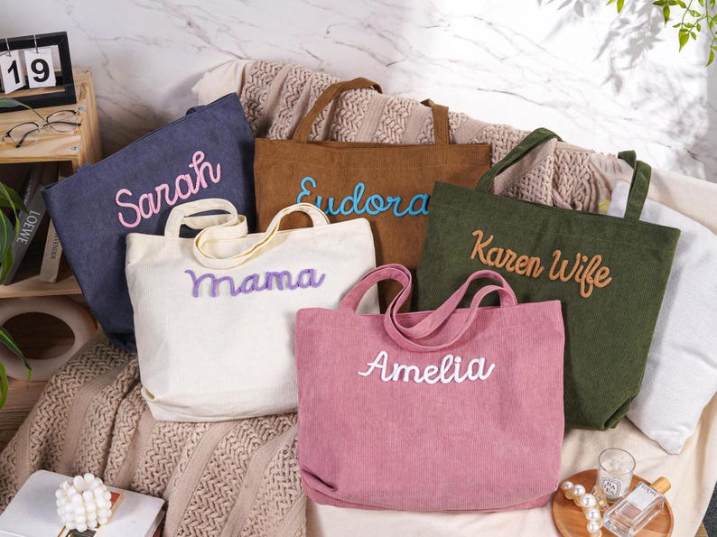 May include: A collection of corduroy tote bags in various colors, including blue, brown, white, green, and pink. Each bag features a different name embroidered in a contrasting color: Sarah, Eudora, Mama, Amelia, and Karen Wife.