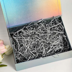 K&ouml;nnte beinhalten: Eine offene, schillernde Geschenkbox mit Klappdeckel, gef&uuml;llt mit silbernem Zerkleinerungspapier. Die Box hat eine metallische Oberfl&auml;che, die die Farben wechselt. Ein Strau&szlig; wei&szlig;er und rosa Tulpen befindet sich in der linken unteren Ecke.