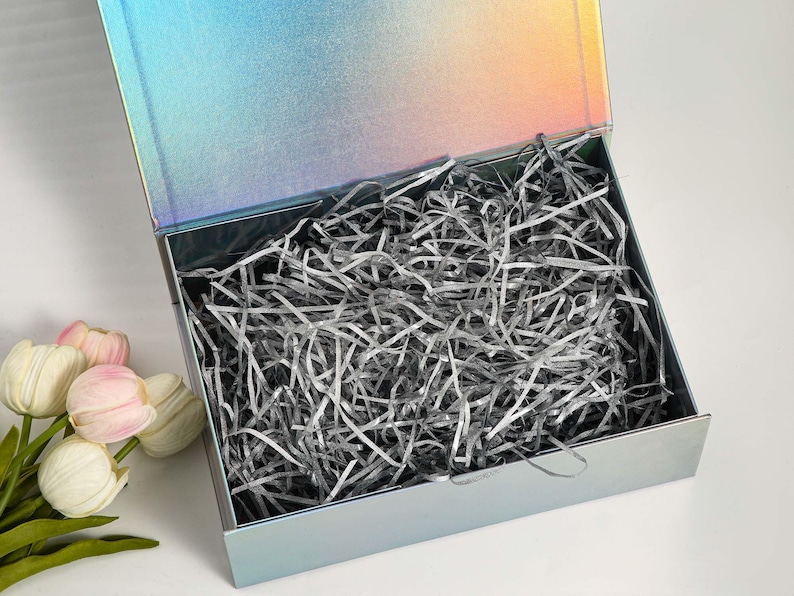 K&ouml;nnte beinhalten: Eine offene, schillernde Geschenkbox mit Klappdeckel, gef&uuml;llt mit silbernem Zerkleinerungspapier. Die Box hat eine metallische Oberfl&auml;che, die die Farben wechselt. Ein Strau&szlig; wei&szlig;er und rosa Tulpen befindet sich in der linken unteren Ecke.