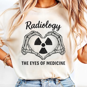Könnte beinhalten: Cremefarbenes T-Shirt mit dem Wort "Radiology" über einer Herzform, die von Skeletthänden gebildet wird. Im Herzen befindet sich ein Strahlungssymbol. Der Text "THE EYES OF MEDICINE" steht darunter.