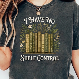 Könnte beinhalten: Dunkelgraues T-Shirt mit dem Text "I Have No Shelf Control". Die Grafik zeigt eine Reihe grüner und beigefarbener Bücher mit floralen und Blattmustern, umgeben von Blumen und Blättern.