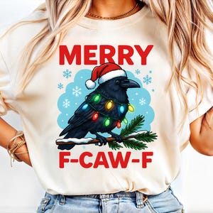 Könnte beinhalten: Cremefarbenes T-Shirt mit einem schwarzen Raben, der einen Weihnachtsmannhut und Lichterketten trägt. Die Worte "MERRY" und "F-CAW-F" sind in Rot über und unter der Rabenillustration gedruckt, mit Schneeflocken auf blauem Hintergrund.