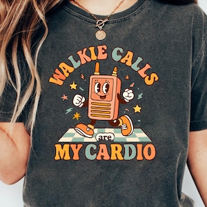 May include: Dark gray t-shirt with a retro design. The graphic features a cartoon walkie-talkie character with the text "WALKIE CALLS are MY CARDIO" in orange, blue, and yellow. The walkie-talkie is on a checkered mat with stars.