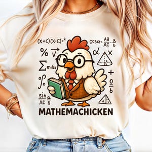 Puede incluir: Camiseta color crema con un dibujo de un pollo de dibujos animados con gafas, corbata y blazer, sosteniendo un libro. La camiseta tiene símbolos matemáticos y el texto "MATHEMACHICKEN".