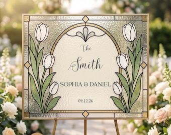 Può includere: Un cartello rettangolare in vetro colorato con cornice dorata che presenta tulipani bianchi con steli e foglie verdi su entrambi i lati. Il centro mostra il testo The Smith, SOPHIA & DANIEL e la data 09.12.26 in caratteri neri su sfondo crema.