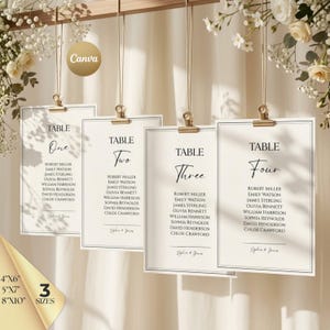 May include: Four rectangular off-white table seating cards with black text, hanging from gold clips. Each card displays a table number, a list of names, and the text Sophie & Jones. The cards are suspended against a light fabric background.