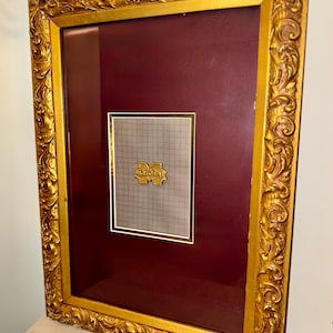 May include: A framed piece featuring a gold-colored, ornate frame. The frame surrounds a burgundy background with a smaller, gold-framed section containing a logo on a grid background. The frame has detailed scrollwork.