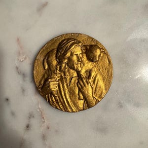 May include: A gold-colored, circular relief sculpture depicting a figure with long hair holding a lamb. The figure is wearing a robe and has a serene expression. The sculpture is likely made of metal and has a textured surface.