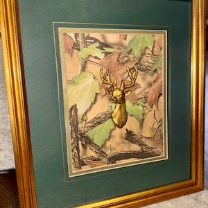May include: Framed artwork featuring a gold-colored deer head mounted on a camouflage background with green and brown leaves. The artwork is set within a green mat and a gold-colored frame, creating a rustic aesthetic.