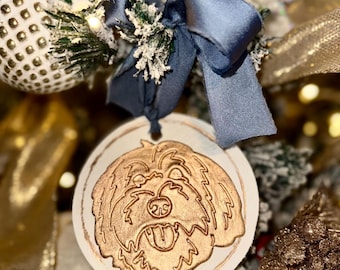 May include: A round, white Christmas ornament with a gold-colored dog design. The ornament is hanging from a navy blue ribbon bow. The background includes a decorated Christmas tree with gold and white accents.