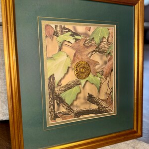 May include: Framed artwork featuring a camouflage design with green and brown leaves, branches, and a gold-colored emblem. The artwork is set within a green mat and a gold-colored wooden frame, creating a decorative piece.