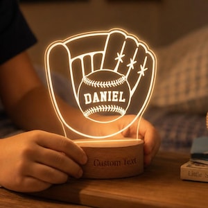 Peut inclure: Une veilleuse en acrylique illuminée en forme de gant de baseball tenant une balle avec le nom DANIEL imprimé sur la balle. La lampe est montée sur une base en bois ronde avec les mots Custom Text gravés sur la face avant.