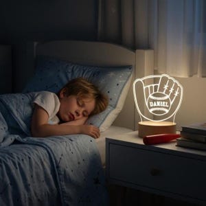 May include: A glowing baseball-themed night light with the name "DANIEL" is on a wooden base, next to a sleeping child. The light is shaped like a baseball glove holding a baseball. The room has a blue comforter and pillow.