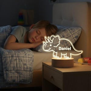 May include: A lit dinosaur-shaped night light with the name "Daniel" in white lettering. The light is on a wooden base, placed on a bedside table next to a sleeping child. The room is dimly lit.