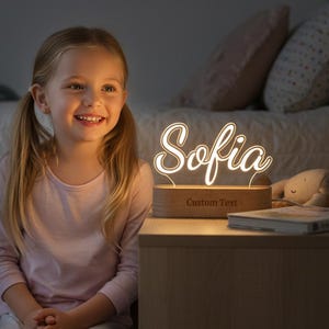 Op de afbeelding: Een oplichtend LED-nachtlampje met de naam "Sofia" in een sierlijk lettertype. Het lampje staat op een houten voet met de tekst "Custom Text". Een boek en een knuffel liggen op het nachtkastje.